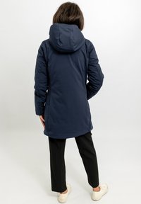 Marineblauwe, gebreide jas met een slank ontwerp, gemaakt van een gladde stof. De achterkant heeft een gebogen zoom en aansluitende mouwen. Draagt over zwarte broeken.