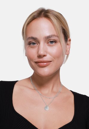 Silberne geschichtete Halskette mit rundem Anhänger, die feine Ketten und kleine Perlenakzente aufweist, getragen mit einem schwarzen gerippten Oberteil.
