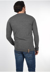 Man met donker haar in een strakke, lange mouwen grijze shirt en blauwe jeans, met de rug naar de camera gekeerd tegen een eenvoudige lichte achtergrond.