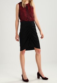 Červený krajkový top bez rukávů ve spojení s černou řasenou sukní, zvýrazněnou detaily na stahování. Doplňují ho bordó lodičky na vysokém podpatku.