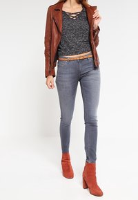 Braune Leder-Bikerjacke über einem grauen strukturierten Oberteil, kombiniert mit schmalen grauen Jeans und roten Wildleder- Stiefeletten. Gürtel mit beigen Akzenten.