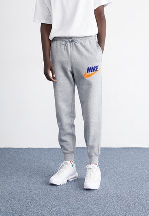 Persoon die lichtgrijze Nike joggingbroek draagt met blauw en oranje logo, witte sneakers en een wit T-shirt, staand op een grijs tapijt.