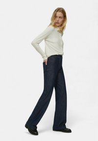 Pantaloni larghi in denim scuro con tasche frontali e cuciture a contrasto, abbinati a una camicia aderente bianca a maniche lunghe con bottoni.