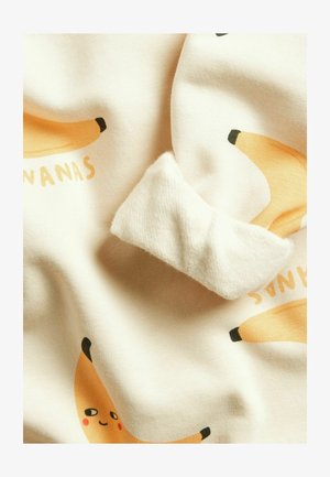 Weicher Stoff mit verspielten Bananenillustrationen, eine Banane hat ein lächelndes Gesicht mit rosigen Wangen auf hellem cremefarbenem Hintergrund.