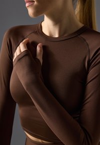 Braunes langärmliges Crop-Top aus glattem, dehnbarem Material. Es hat einen hohen Halsausschnitt und eine figurbetonte Passform, mit Daumenlöchern in den Ärmeln.