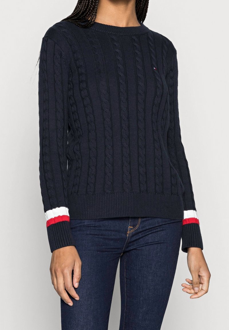 Pull en maille torsadée bleu marine avec un col rond et un ourlet côtelé. Présente des accents rayés rouge, blanc et bleu sur les manches.