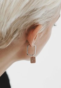 Pendientes de metal que presentan un aro rectangular de plata y un pequeño charm cuadrado de oro rosa con una letra "IV" embosada. Textura suave.