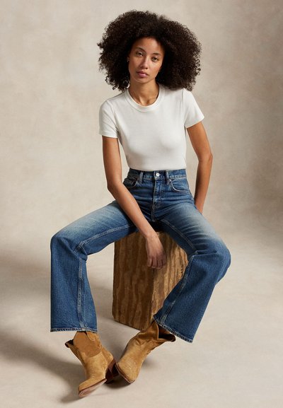 Žena s kudrnatými vlasy sedí na dřevěném bloku, má na sobě bílou košili s krátkým rukávem, modré džíny s vysokým pasem a hnědé semišové boty.