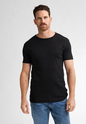 Man met kort krullend haar en baard, gekleed in een zwart T-shirt met korte mouwen en een blauwe spijkerbroek, staand tegen een effen grijze achtergrond.