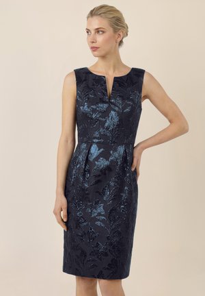 Frau mit blonden Haaren in einem ärmellosen, dunkelblauen, knielangen Kleid aus Brokat mit Blumenmuster, die mit einer Hand in der Hüfte steht.