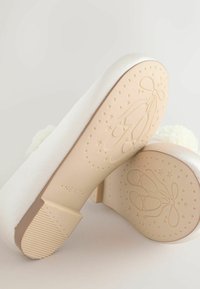 Bílé baletní baleríny s texturovaným plátěným svrškem a květinovým akcentem, které mají béžovou gumovou podrážku s reliéfem hvězdiček a baletních bot.