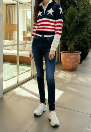 Femme portant un pull à étoiles et rayures, un jean bleu foncé et des baskets blanches, debout à l'intérieur près d'une porte en verre et d'une grande plante en pot.