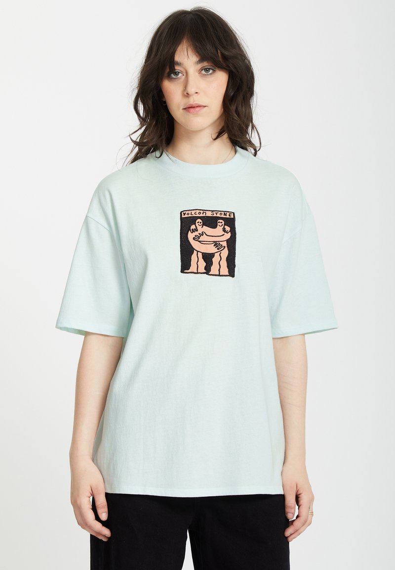 Helles blaues Baumwoll-T-Shirt mit oversized Schnitt. Mit einem zentralen bestickten Design, das zwei Figuren zeigt. Kurze Ärmel und runder Halsausschnitt.