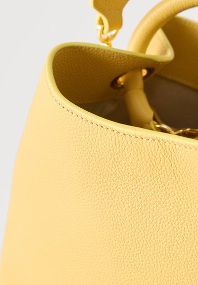 Κίτρινη τσάντα χειρός από δέρμα με υφή, ορατές ραφές, στρογγυλές λαβές και μεταλλικά στοιχεία σε χρυσό χρώμα σε ανοιχτό φόντο.