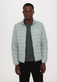 Light gray padded jacket with a high collar, featuring horizontal stitch lines and a logo on the chest, worn over a dark shirt.