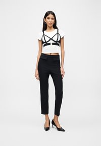 Γυναίκα που φοράει λευκό κοντό t-shirt με μαύρο ιμάντα harness από πάνω, μαύρο tailored παντελόνι και μαύρες ψηλοτάκουνες γόβες, στέκεται μπροστά σε λευκό φόντο.