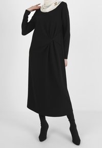 Vestido negro de manga larga con detalle de nudo retorcido en la cintura, hecho de una tela suave. Accesorado con una bufanda de color claro.