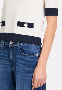 Korte mouw, gebreide top in off-white met marineblauwe bies. Heeft twee marineblauwe zakken met zilveren knopen. Gecombineerd met middelmatige wassing jeans.