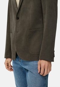 Brun fløjl blazer med en enkelt knap, stikkraver og to fremadskuende lommer; parret med lyseblå jeans. Detaljer af tekstur er synlige.