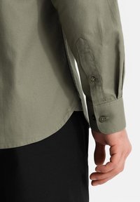 Camicia a maniche lunghe di colore verde oliva, realizzata in tessuto testurizzato, con polsini con bottoni e orlo curvo, abbinata a pantaloni neri.