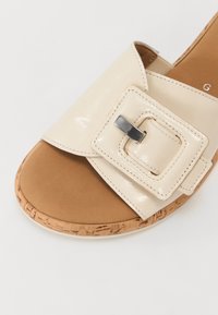 Sandale en cuir verni crème avec une grande boucle carrée, une semelle intérieure rembourrée couleur tan et un bord de semelle texturé en liège.