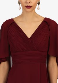 Bordeauxfarbenes Kleid mit plissiertem Stoff, V-Ausschnitt und schwingenden Ärmeln. Goldene Ohrringe mit Perlenakzenten vervollständigen den Look.