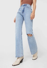 Světle modré džínové kalhoty s vysokým pasem a volným střihem, které mají velkou díru na koleni a neupravené okraje. Kombinované s béžovými botami na podpatku.