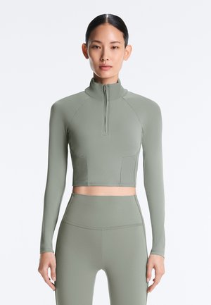 Mlada ženska v usklajeni svetlo zeleni kratki zgornji majici z dolgimi rokavi in legicah z visokim pasom, stoji pred enobarvno ozadjem.