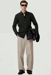 Camicetta nera a maniche lunghe con bottoni, pantaloni beige a gamba larga, che tiene una borsa nera e indossa occhiali da sole. Focalizzazione su linee semplici e pulite.