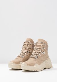 Botas de tobillo de ante beige con una suela gruesa de color crema, que presentan ojales plateados, cordones gruesos y un acolchado en el cuello para mayor soporte.