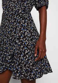 Robe noire à fleurs avec un ourlet volanté, un petit motif de fleurs bleues et blanches, des manches bouffantes et une taille cintrée. Détails autour de l'encolure.