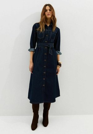 Kobieta ubrana w ciemną jeansową sukienkę midi zapinaną na guziki, z podwiniętymi rękawami i paskiem, do tego ciemnobrązowe kozaki za kolano oraz bransoletki.