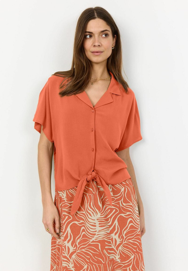 Blusa arancione a maniche corte con colletto, caratterizzata da un dettaglio di fiocco frontale, abbinata a una gonna stampata in tonalità complementari.