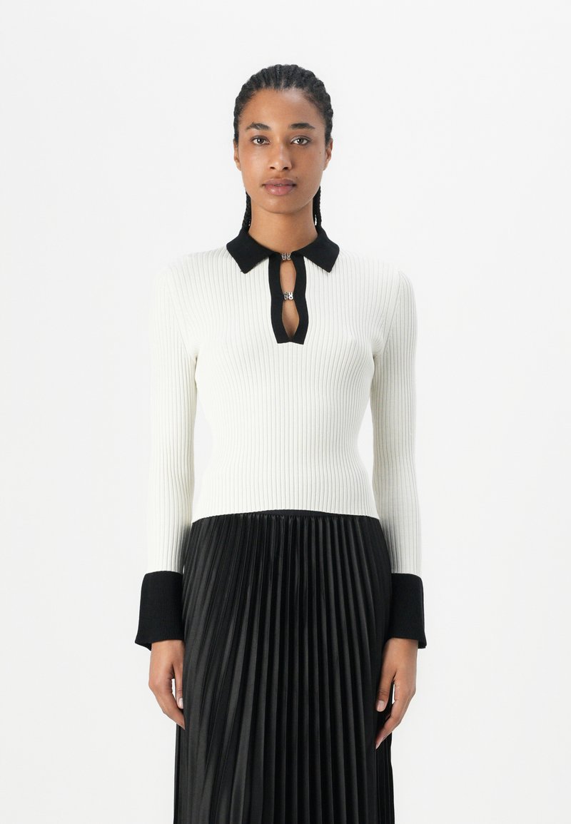 White ribbed long-sleeve top with a black collar, keyhole neckline, and black cuffs, paired with a black pleated skirt.