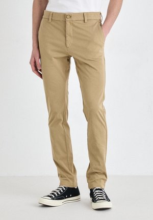 Personne portant un pantalon beige slim et des baskets noires basses avec des lacets blancs, debout devant un fond blanc.