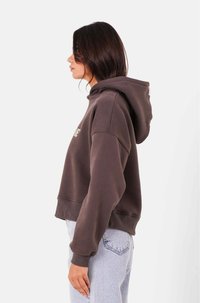 Sudadera corta marrón con capucha ajustable, puños acanalados y un logo bordado en la parte frontal. Combinada con unos jeans de color azul claro.