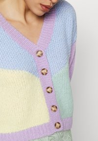 Wielokolorowy sweter z dzianiny z pastelowymi odcieniami: jasny fiolet, żółty i miętowy. Ma dekolt w serek i duże brązowe guziki.