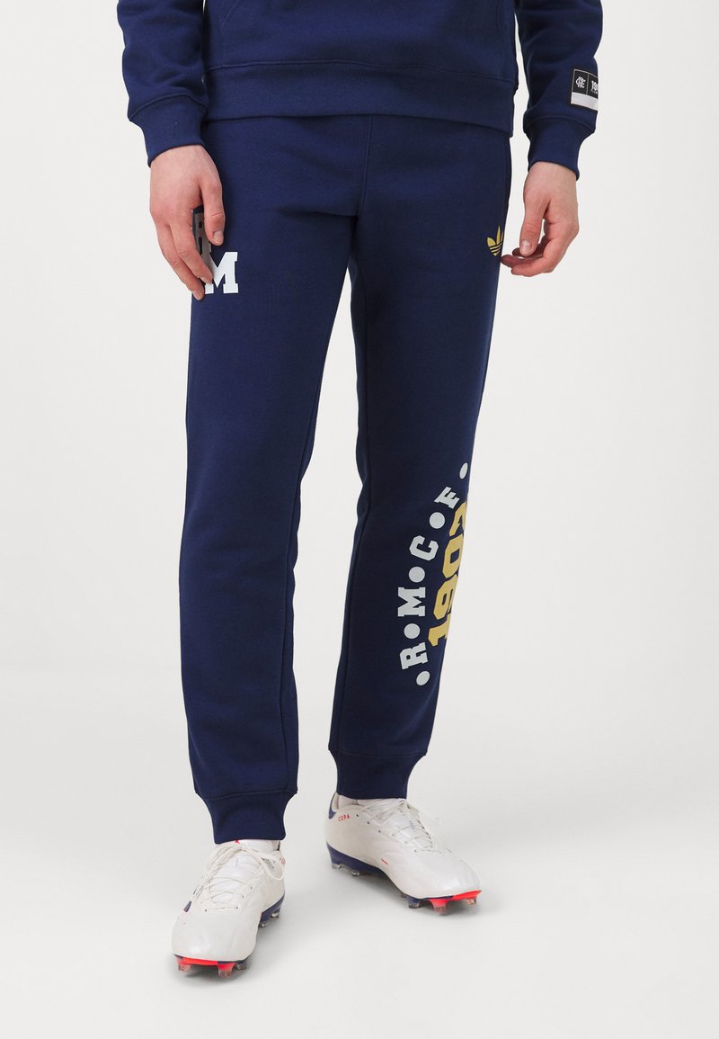 Calças de fato de cor azul marinho, feitas de um tecido macio, apresentam a letra branca "M" e as impressões amarelas "R.M.C.F. 1903" ao longo da perna, com bainhas ajustadas.