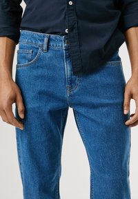 Kék farmernadrág egyenes szabással, klasszikus öt zsebes kialakítással, kiemelő varrással és gombos záródással.