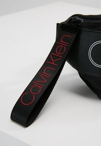 Banane noire en matière texturée avec un logo circulaire, dotée d'une sangle embossée du nom « Calvin Klein » en lettres rouges. Fermeture à glissière.