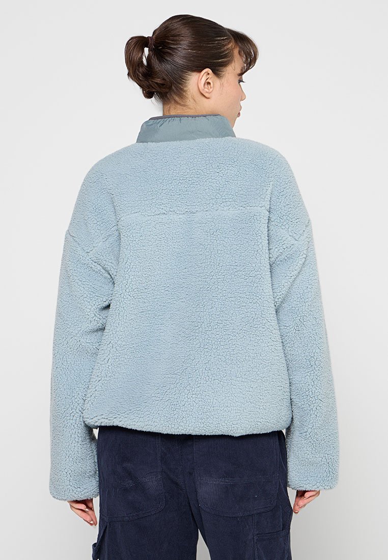 Hellblaue Fleecejacke mit einer strukturierten, kuscheligen Außenfläche und einem kontrastierenden, weichen Kragen. Sie hat einen lockeren Schnitt und lange Ärmel.