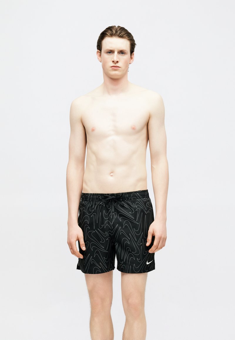 Άνδρας χωρίς πουκάμισο, φορώντας μαύρα μαγιό με αφηρημένα λευκά σχέδια γραμμών και λευκό λογότυπο της Nike, στέκεται μπροστά σε ένα απλό φόντο.