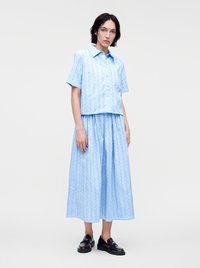 Femme debout portant une chemise bleu clair à manches courtes boutonnée et une jupe longue assortie avec un motif œillet ainsi que des mocassins en cuir noir.