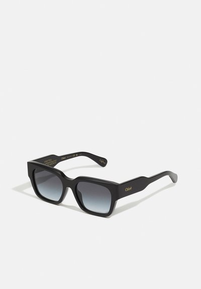 Lunettes de soleil en acétate noir avec une monture carrée audacieuse, des verres dégradés et un logo doré à l'intérieur des branches. Présente une finition texturée.