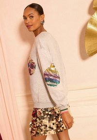 Sudadera gris con acentos de lentejuelas en las mangas, combinada con una falda ajustada cubierta de lentejuelas doradas. Detalles estilizados, texturizados y coloridos a lo largo.