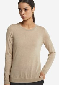 Beige långärmad tröja med rund halsringning, mjukt tyg och ribbade muddar samt nederkant; bärs med modellens hand vilande på höften.