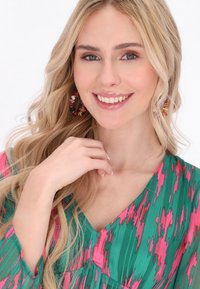 Bluză cu model verde și roz, cu decolteu în V, dintr-un material moale, cu mâneci lungi și cercei cu mărgele. Textură catifelată și culori vibrante.