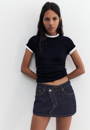 Frau mit langen dunklen Haaren trägt ein eng anliegendes schwarzes T-Shirt mit weißem Rand und einen dunklen Jeans-Minirock, steht vor weißem Hintergrund.