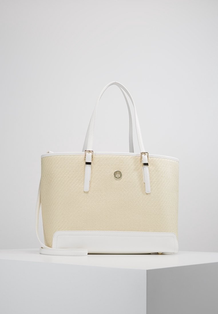 Sac cabas beige en tissu tissé avec accents en cuir blanc et doubles poignées. Dispose d'un logo en métal et d'une sangle amovible. Texture lisse.