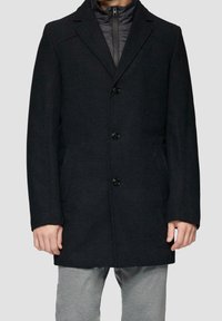 Man in een donker gestructureerd jasje met drie zwarte knopen over een zwart ritsjack en lichtgrijze broek, staande tegen een effen achtergrond.